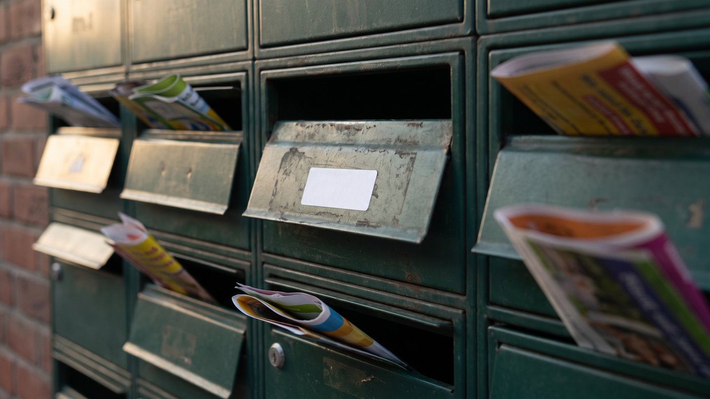 Deux rangées de boîtes aux lettres vertes d'immeuble, l'un des volets portant une étiquette blanche vierge, les autres fentes garnies de prospectus colorés